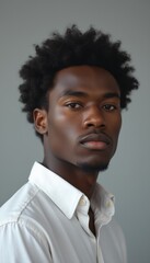 Obraz premium A striking portrait of a young Black man with a confident expression, showcasing his natural curly hair against a soft gray background. His poised demeanor and stylish white shirt exude elegance and