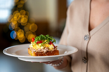 A close-up of a delicious open-faced sandwich topped with scrambled eggs, tuna, cherry tomatoes, and fresh greens. A festive bokeh background adds a warm atmosphere to the scene.
