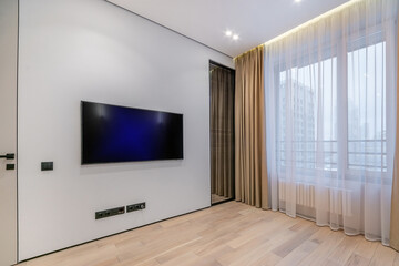 The living room in the apartment has a large window and an installed TV, unfurnished. .White room...