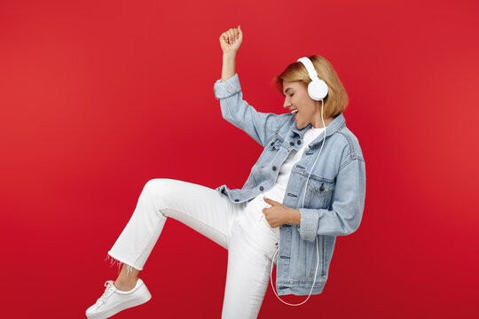 Stylish young blonde woman in casual white outfit listening music in headphones over red background.