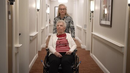 A daughter pushes her elderly mother in a wheelchair down a well-lit corridor of a care facility, sharing a meaningful moment together.