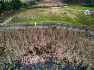 Person in Yellow Protective Gear on a Wooden Boardwalk Over a Marsh. High-angle view of a person wearing a bright yellow protective suit walking across a wooden boardwalk over a marsh area. Sakarya