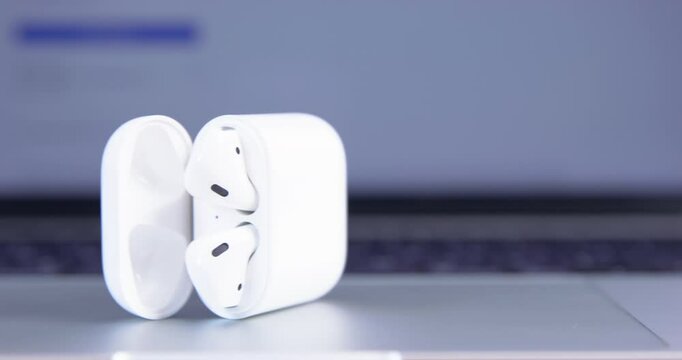 Stylish wireless headphones lie in open white smooth original case on operating grey laptop.