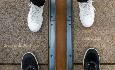 High-angle shot of two people standing between the Greenwich Prime Meridian marked by the brass strip on the ground in the courtyard of the Royal Observatory with markers of well known capital cities.