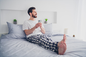 Photo of funky happy mature guy sleepwear pajama sitting bed texting modern gadget indoors home...