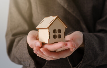 A person holds a small white model house with windows and a door in their hands, symbolizing home ownership and real estate investment.