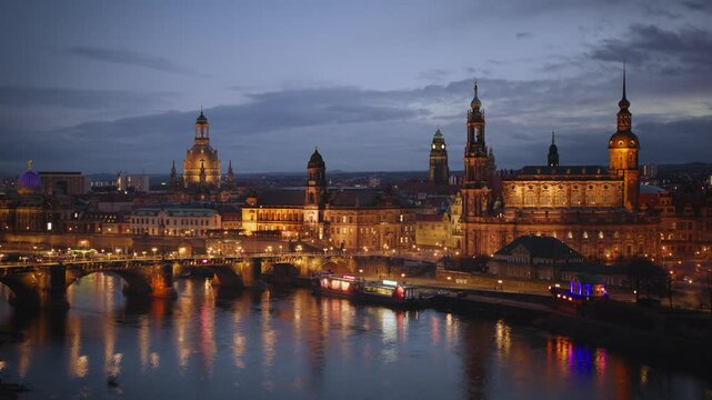 Aerial view of Dresden's historic Old Town at night, with the Elbe River reflecting city lights. The drone glides above the river, showcasing illuminated landmarks and bridges. Drone flies forward.