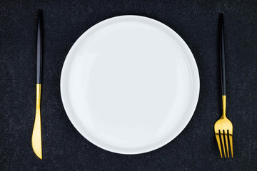 Elegant white plate with black and gold utensils arranged on a dark textured surface for a refined dining experience