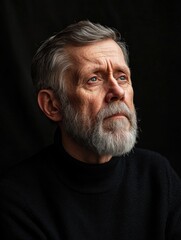 Obraz premium Reflective elderly man with gray beard in thoughtful pose wearing black sweater against rich black background, captured in soft natural lighting.