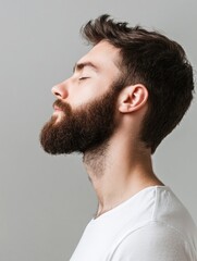 Obraz premium Profile of a handsome young man with a beard, eyes closed, set against a soft grey background, natural lighting highlighting facial features, casual white shirt.
