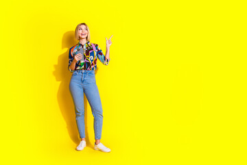 Full size photo of pretty young girl hold device point look empty space wear trendy colorful outfit isolated on yellow color background