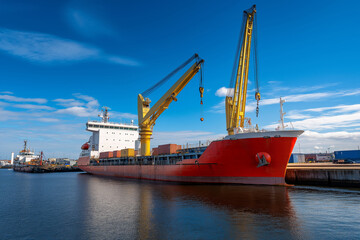 Fototapeta premium Large cargo ship with containers and yellow crane docked at a port under a bright blue sky with calm waters..