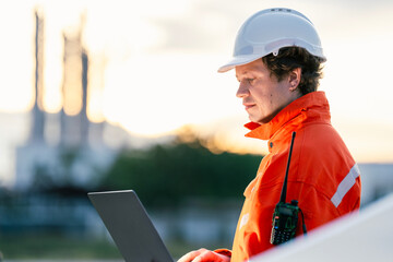 Engineer in bright orange reflective suit with safety helmet and laptop, standing in front of an...