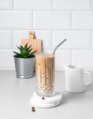 Cold latte coffee in a tall glass with milk and straw on a light table on a white brick background
