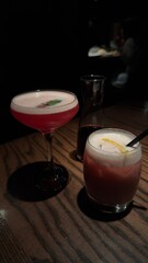 Two vibrant cocktails on a wooden table in dim lighting: a red drink in a coupe glass with a mint garnish, and a frothy cocktail in a short glass with a lemon peel. Cozy, intimate ambiance.