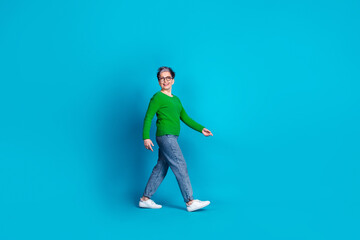 Confident elderly woman walking with a smile, wearing glasses and green sweater against a blue...