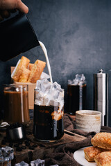 Hand of barista pours refreshing Milk on iced coffee into a glass to make ice latte , capturing the perfect vertical shot with crispy snacks for coffee lovers.