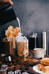 Hand of barista pours refreshing Milk on iced coffee into a glass to make ice latte , capturing the perfect vertical shot with crispy snacks for coffee lovers.