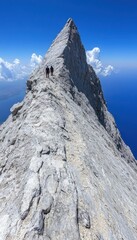 Hikers ascend a dramatic, rocky mountain ridge against a vibrant blue sky and ocean backdrop.