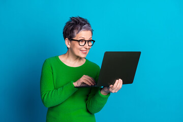 Mature woman with gray hair using laptop in front of vibrant blue background, wearing glasses and green sweater