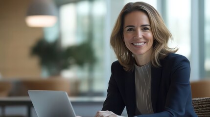 A confident businesswoman in a modern office, smiling at the camera while using a laptop, showing teamwork and professionalism