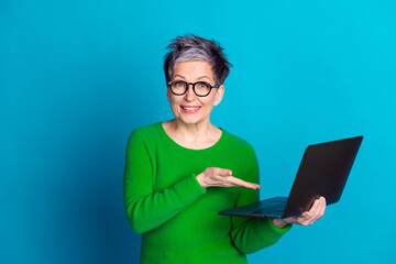 Elderly woman in glasses showcasing technology with laptop against blue background