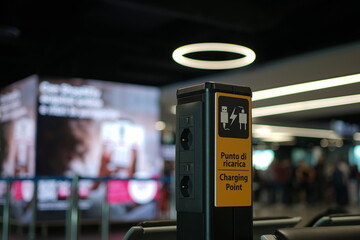 he charging station, with several sockets and USB ports, marked as "Charging Point" in Italian and English, is located in a modern, stylish airport building.