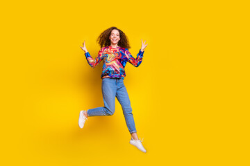 Joyful young woman with curly red hair wearing trendy colorful jumper jumps against a bright yellow background