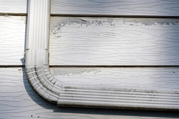 Closeup of an old downspout as it bends along the side of a house with peeling paint on the aluminum siding
