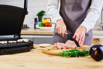Preparation on electric grill. Woman in white shirt and apron standing and cutting meat on the kitchen. Electric grill barbecue.