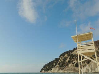 observation tower on empty beach made of light wood with red and white flag showing strong wind against background of cliff on seashore on sunny day