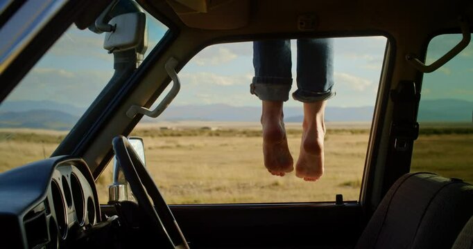 A tranquil and serene outdoor scene features female feet playfully dangling from a car window amidst a picturesque landscape