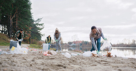 Group of people walking on beach of local lake and picking up trash left by others. Volunteers...