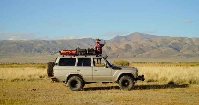 Young woman person stands proudly on an impressive offroad vehicle and take selfy on phone on roof a vehicle, surrounded by stunning mountains and open plains