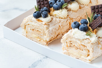 Homemade meringue roll decorated with almond flakes, blueberries and mini chocolate bars in the white gift box on the white background. Meringue roulade with whipped cream on top.
