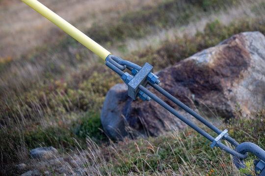 A guy wire secured to the ground from a distribution utility pole. The galvanized wire is wrapped in a yellow insulator covering. The anchor and tensioned stability cables are connected to a hook. 