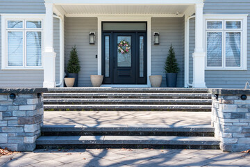The entrance to a large luxury grey wooden house with white trim. The black door has side windows....