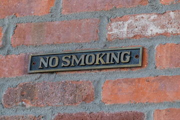 A brass sign with no smoking hanging on the exterior of a red brick wall. The background of the...