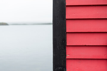 A vibrant pink wooden exterior wall cladded with pink clapboard siding. The fishing shed's trim is black wood. The water in the cove is smooth and it's a grey foggy day. There's land in the distance.