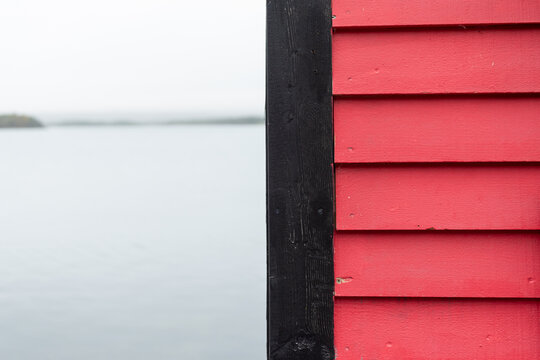 A vibrant pink wooden exterior wall cladded with pink clapboard siding. The fishing shed's trim is black wood. The water in the cove is smooth and it's a grey foggy day. There's land in the distance.