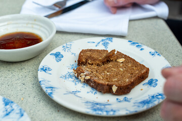 A man tears a small slice of pumpernickel bread on a white and blue China plate. There's a balsamic vinaigrette dipping sauce in a small white dish on the table next to cutlery and a white napkin. 