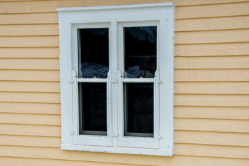 Fototapeta premium The exterior of a yellow wooden cottage with an antique glass double hung window. There's white wide wooden trim around the vintage window. The inside of the building is dark. The siding is pine board