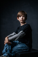 Studio portrait of a young boy sitting against a dark background, wearing casual clothes. The soft lighting highlights his contemplative expression.