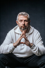 A close-up studio portrait of a mature man with salt-and-pepper hair and a serious, thoughtful expression. Wearing a white hoodie, he poses with hands clasped in front of him