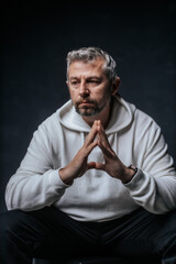 A close-up studio portrait of a mature man with salt-and-pepper hair and a serious, thoughtful expression. Wearing a white hoodie, he poses with hands clasped in front of him