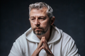 A close-up studio portrait of a mature man with salt-and-pepper hair and a serious, thoughtful expression. Wearing a white hoodie, he poses with hands clasped in front of him