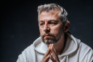 A close-up studio portrait of a mature man with salt-and-pepper hair and a serious, thoughtful expression. Wearing a white hoodie, he poses with hands clasped in front of him