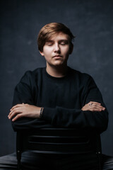 A young man in a black sweater sits with arms crossed, gazing thoughtfully into the distance. The...