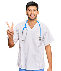 Fototapeta premium Young handsome man wearing doctor uniform and stethoscope smiling with happy face winking at the camera doing victory sign. number two.