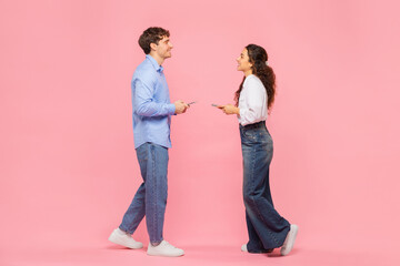 Full length portrait of positive European couple using cellphones, walking to each other and smiling isolated over pink studio background